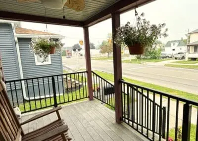Covered front porch with wooden rocking chairs, black railing, hanging flower baskets, and a leaf-shaped ceiling fan, built by Wayne Craft